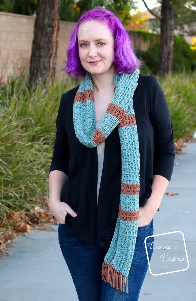 [Image description} A white woman with purple hair models the Adair Scarf wrapped around her neck and one end back over her shoulder, while standing on a sidewalk in front of trees and bushes 