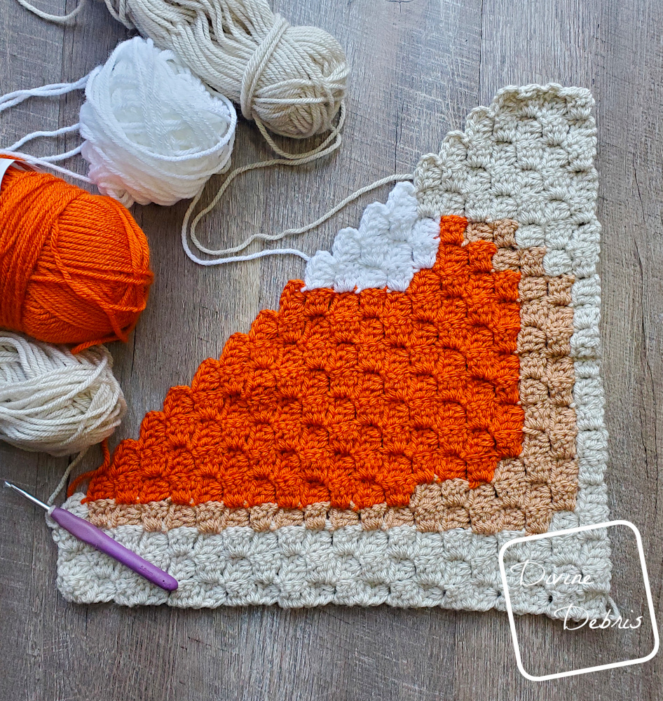 [Image description] Top down view of a half- finished C2C Pumpkin Pie Square laying on a wood grain background, with skeins of yarn around the top left edge of the square.
