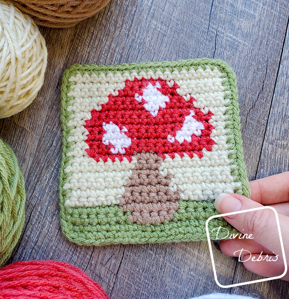 [Image description] Top down view of the Cute Mushroom Coaster crochet pattern, with cakes of yarn arranged around the left side and a white woman's hand holding the bottom right corner.