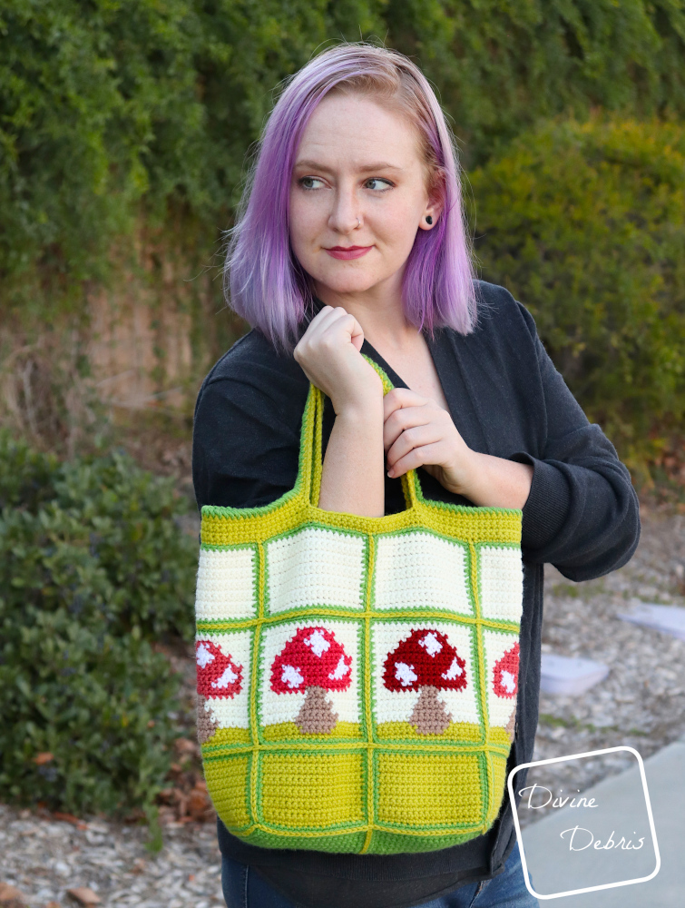 [image description] a white woman stands looking to the side in front of a ivy covered wall holding the Cute Mushrooms Bag to the camera