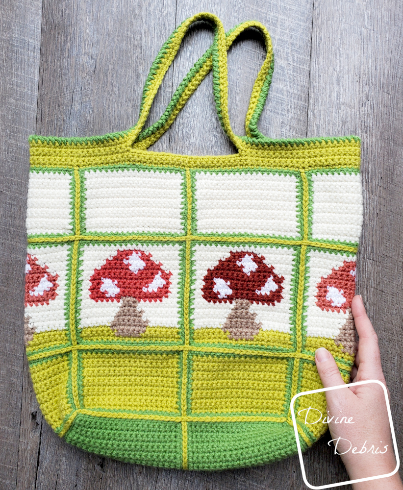 [Image description] Top down view of the Cute Mushrooms Bag crochet pattern on a wood grain background with a white woman's hand holding the bottom right corner