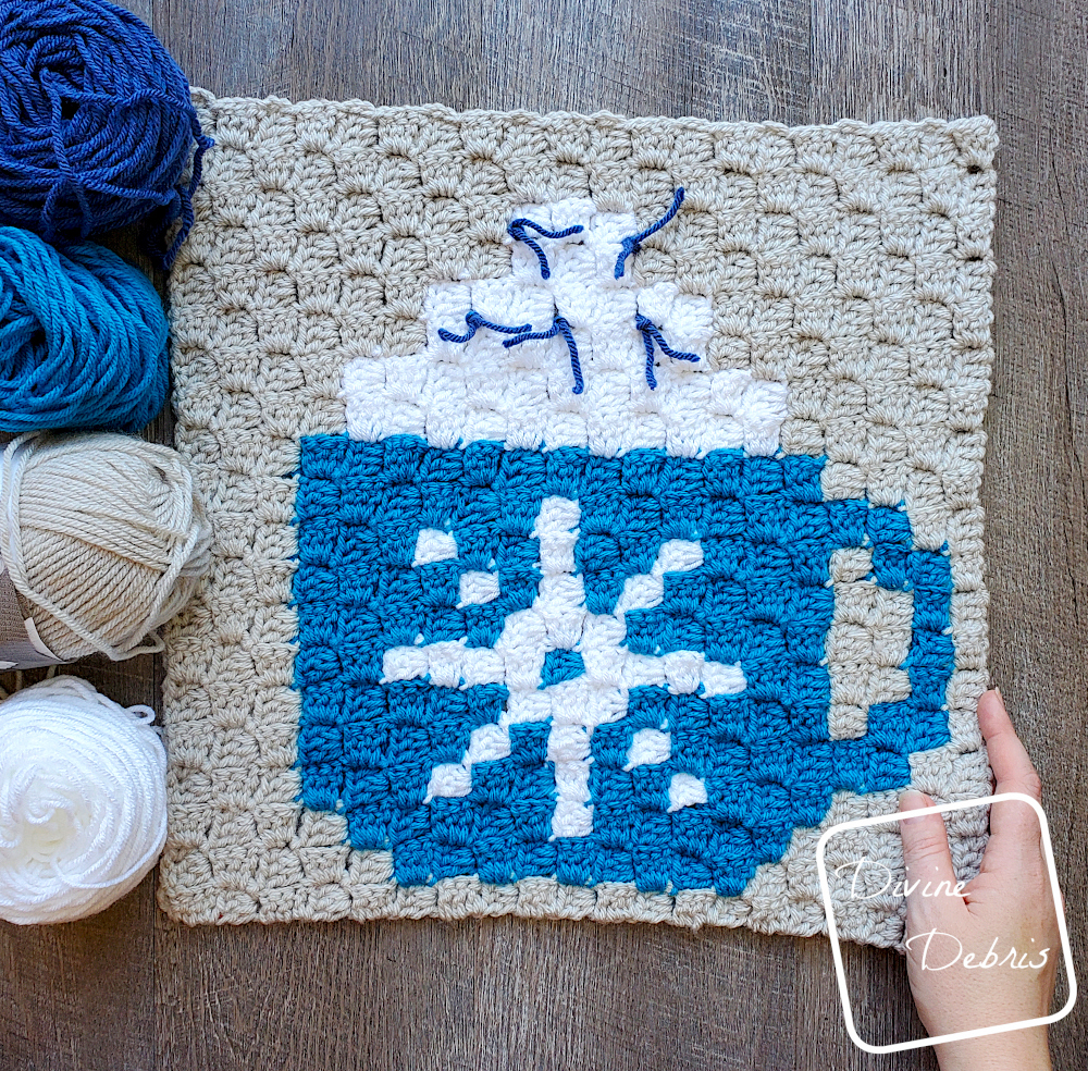 [Image description] Top down view of a C2C Cup of Cocoa Afghan Square laying on a wood grain background, with skeins of yarn around the top left edge of the square and a white woman's hand holding the bottom right corner of the square.