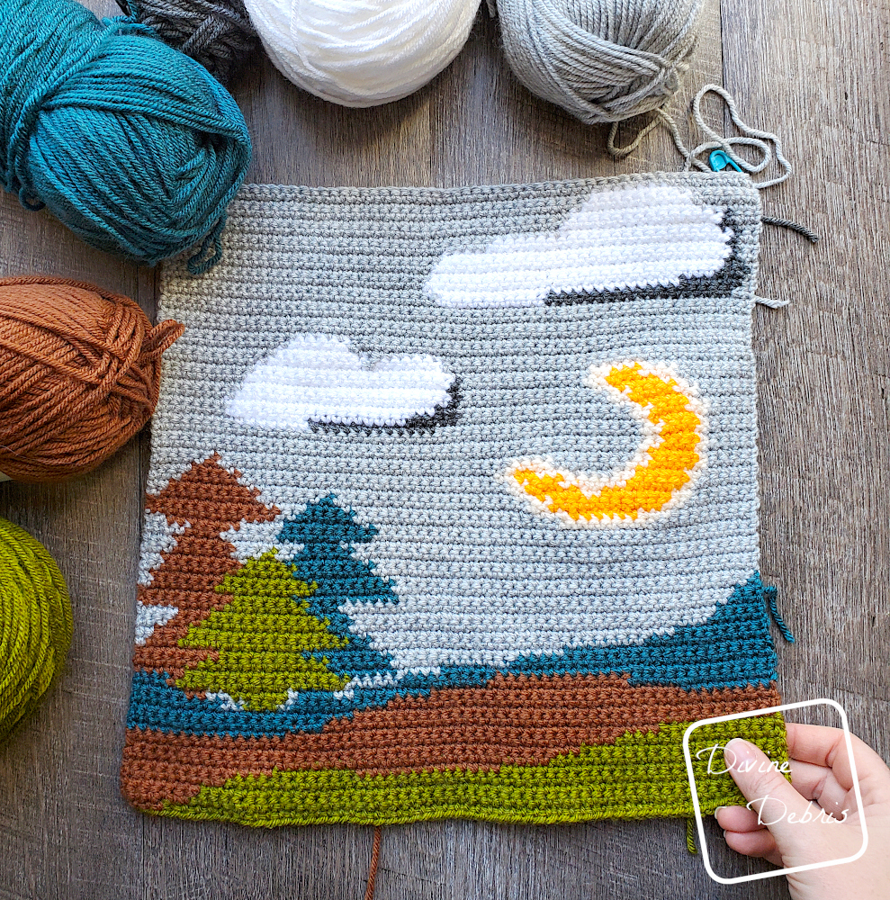[Image description] A top down look at a unfinished Moonlight Trees Wall Hanging laying on a wood grain background, with 5 skeins/ cakes of yarn on the left side of the wall hanging and a white woman's hand holding the bottom right corner.