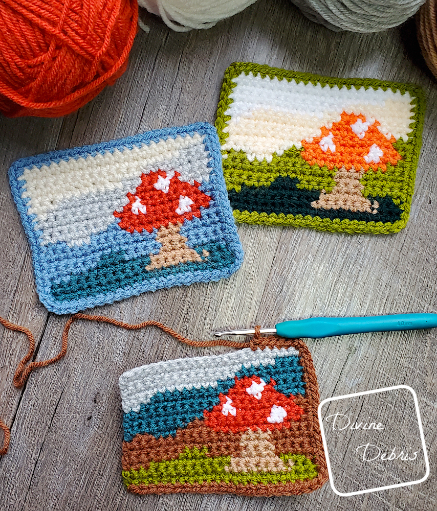 [Image description] Top down view of 3 Mushroom Landscape Mug Rugs, one unfinished, on a wood grain background, 2 skeins of yarn can be seen at the top of the photo