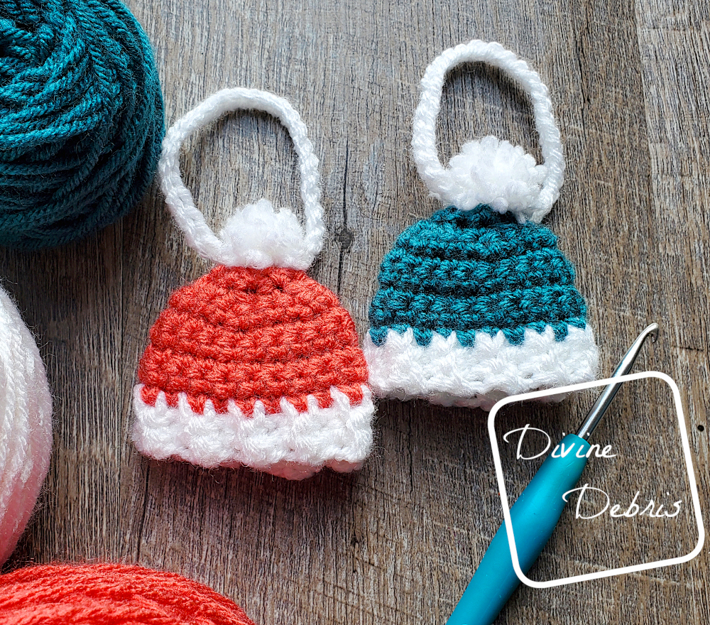 [Image description] Top down flat lay of 2 Winter Beanie Ornament, one red one blue, on a wood grain background with cakes of yarn around the left side and a blue crochet hook on the bottom right