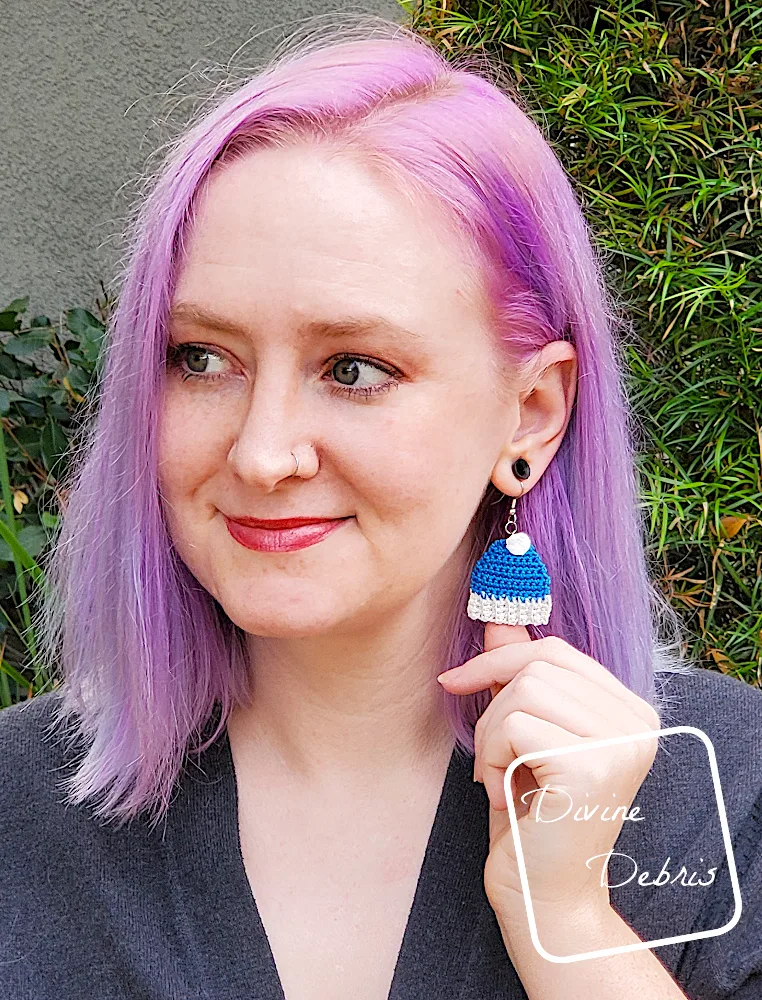 [Image description] A white woman with purple hair stands in front of a green bush looking to the side, holding the bottom of a blue Winter Beanie Ornament Earring toward the camera.