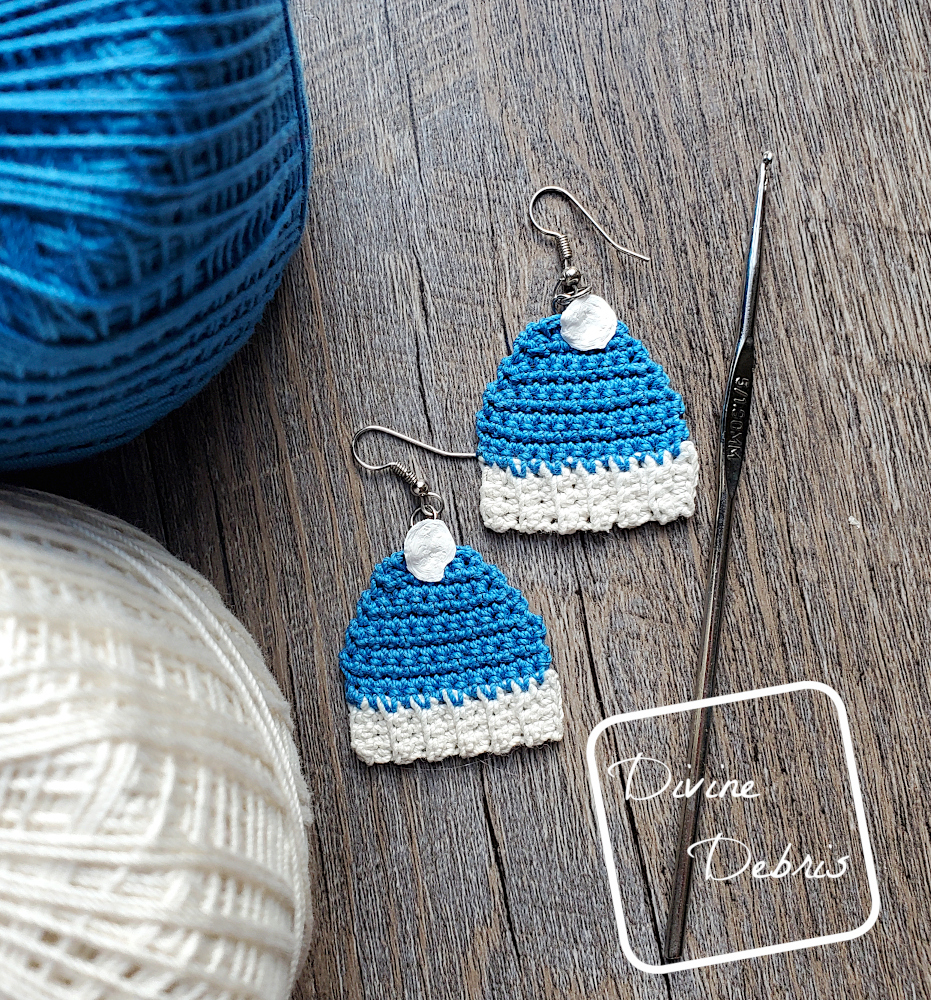 [Image description] Top down view of the Winter Beanie Ornament Earrings in blue on a wood grain background. A crochet hook sits on the right and cakes of crochet thread on the left.