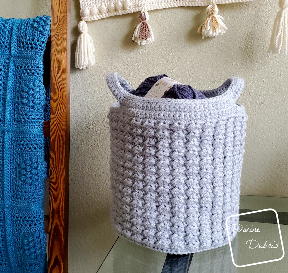 [image description] the Lucinda Basket crochet pattern made in light blue yarn sits on a glass table in front of a tan wall with a blanket holder on the left side of the photo.