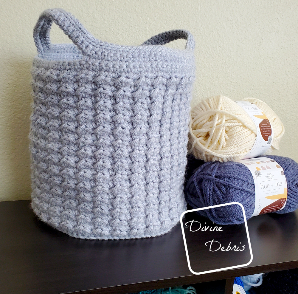 [image description] the Lucinda Basket crochet pattern made in light blue yarn sits in front  of a tan wall with skeins of yarn sitting next to it.