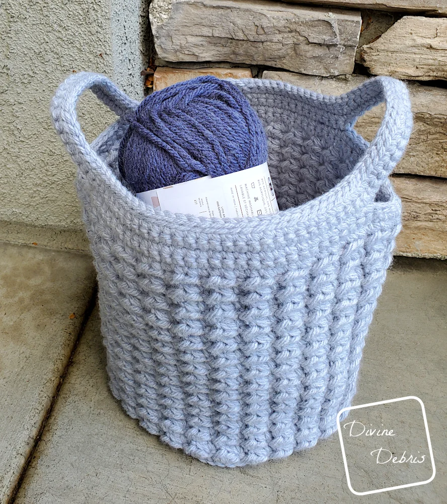 [image description] a top down view of the Lucinda Basket crochet pattern made in light blue yarn sitting in front of stone wall.
