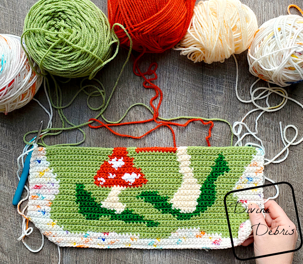 [Image description] Top down view of the unfinished Mushroom Love Wall Hanging laying on a wood grain background with 5 cakes of yarn still attached and along the top of the photo and a white woman's hand holding the bottom right corner.