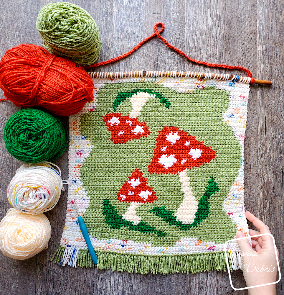 [Image description] Top down view of the Mushroom Love Wall Hanging laying on a wood grain background with 5 cakes of yarn to the left of the wall hanging and a white woman's hand holding the bottom right corner,