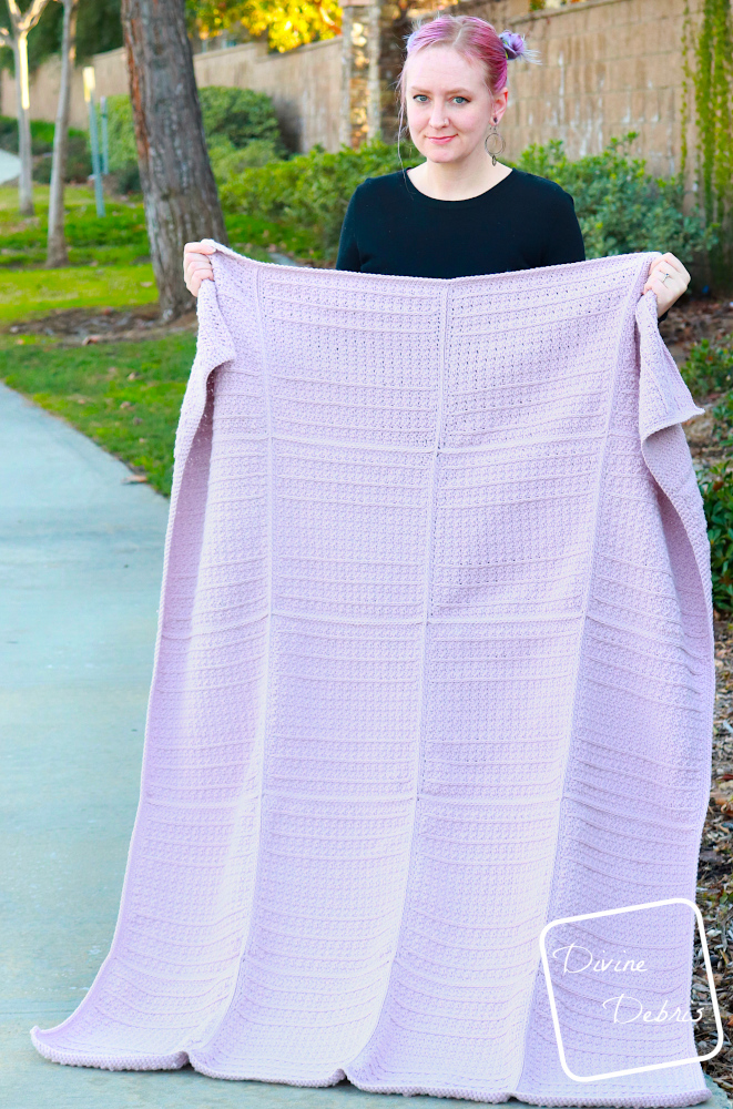 [Image description] A white woman in a black sweater with purple hair stands in front of a tree and grass holding the lavender Kieran Blanket out wide