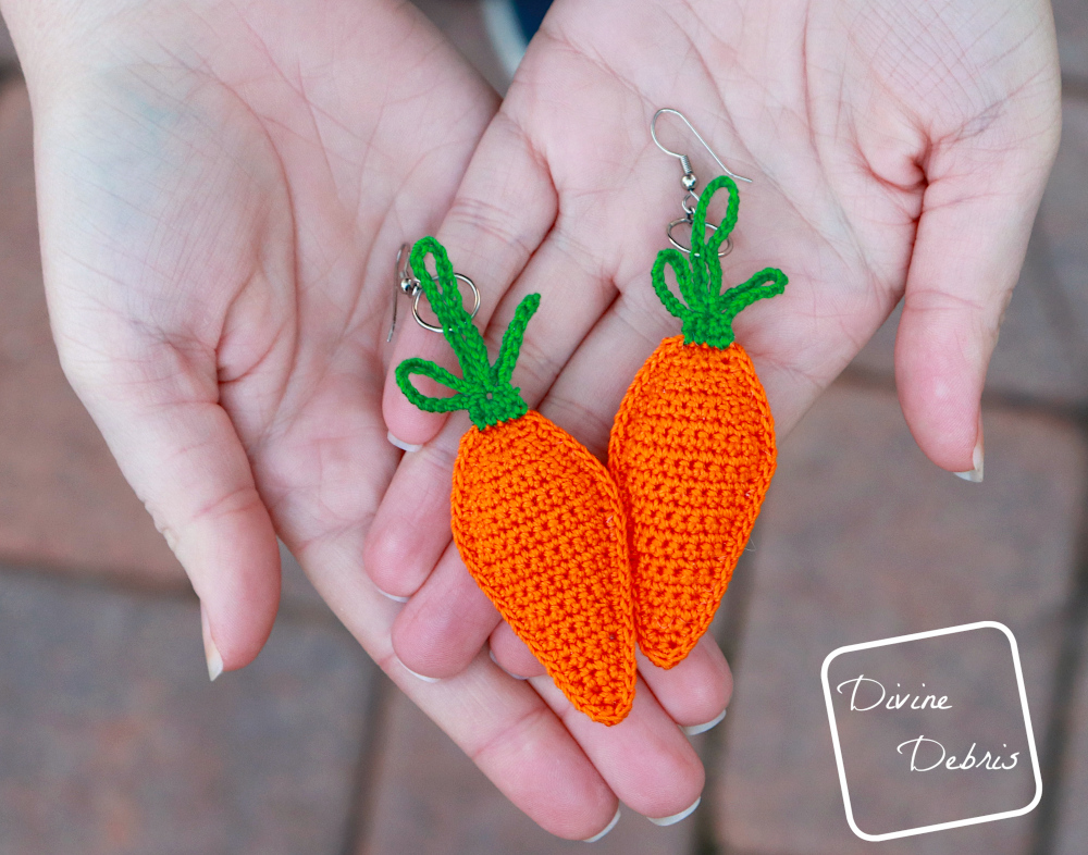 [Image description] A top down look on a pair of white woman's hands holding a pair of Stuffed Carrots Crochet Earrings