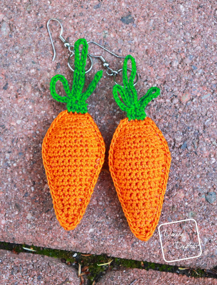 [Image description] 2 Finished Stuffed Carrots Crochet Earrings on a red stone background
