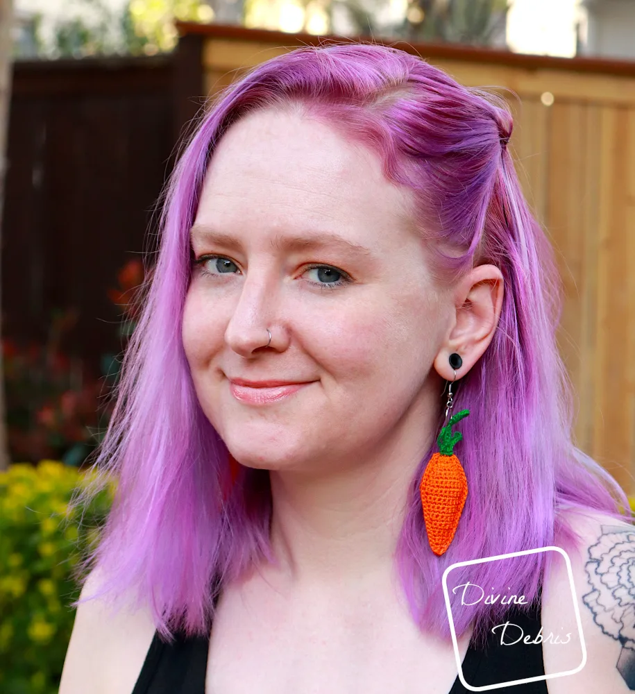 [Image description] A white woman with purple hair stands in front of a tan fence holding her Stuffed Carrots Crochet Earrings toward the camera.