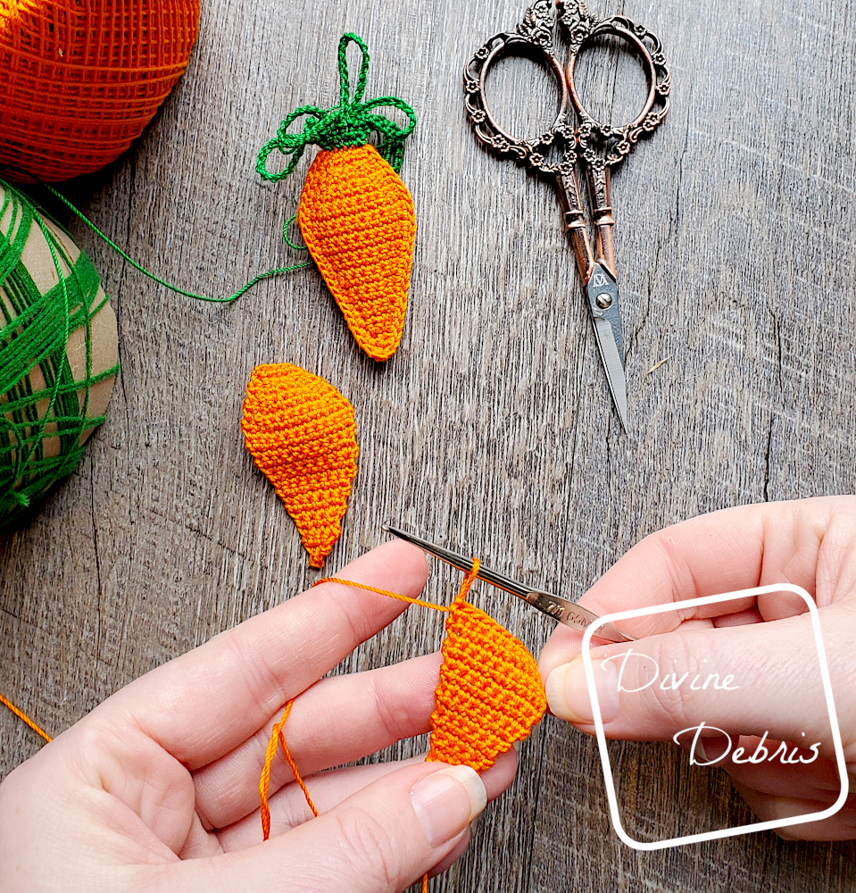 [Image description] Top down look onto a white woman's hands making one of the carrots in the Stuffed Carrots Earrings. A finished Carrot Earring lays on a wood grain background with a pair of scissors to the right and skeins of crochet thread to the left.