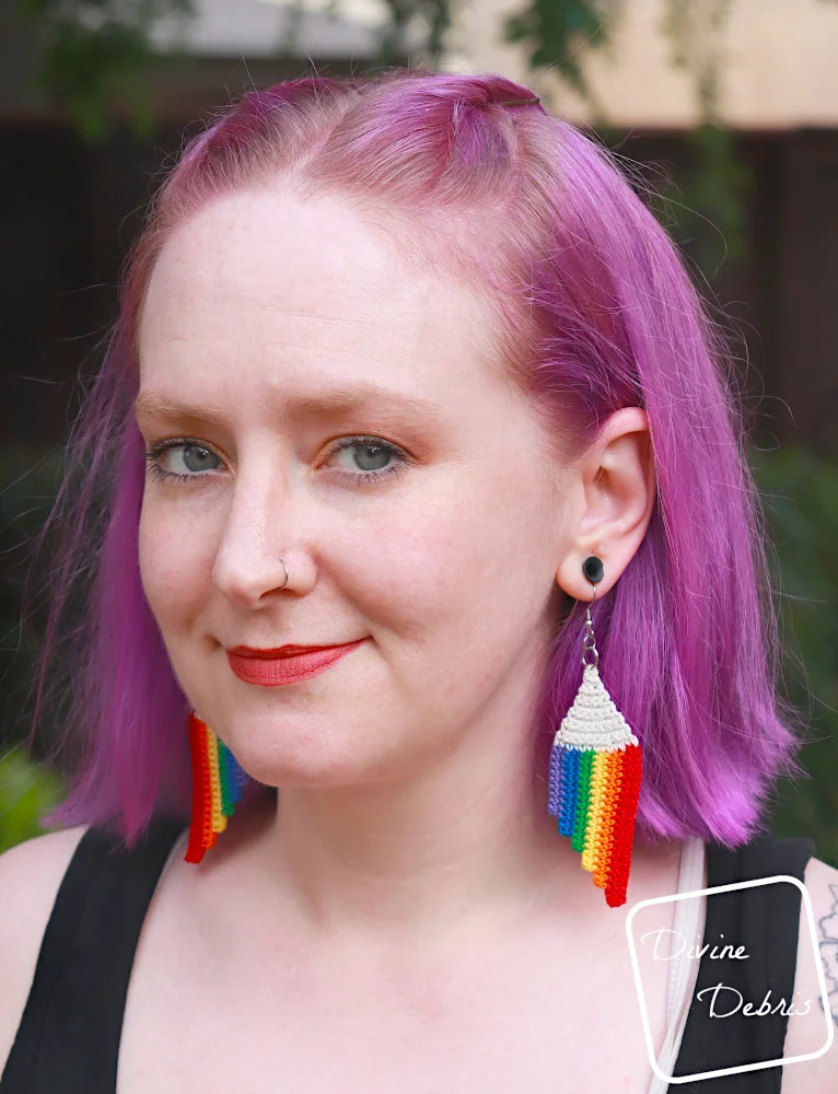 [Image description] A white woman with purple hair stands facing the camera wearing the Cascade Rainbow Earrings crochet pattern.