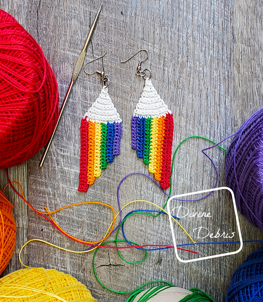 [Image description] Top down view of the Cascade Rainbow Earrings crochet pattern laying on fake wood background with skeins of crochet thread and strands around the earrings.
