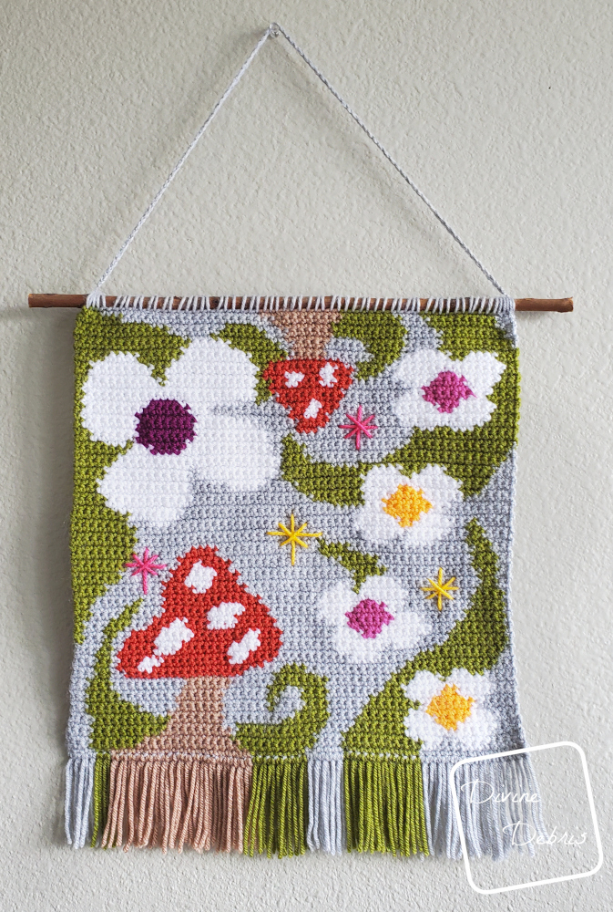 [Image description] A colorful mushroom and white flower wall hanging, the 'Shrooms and Blooms Wall Hanging, hangs against a tan wall