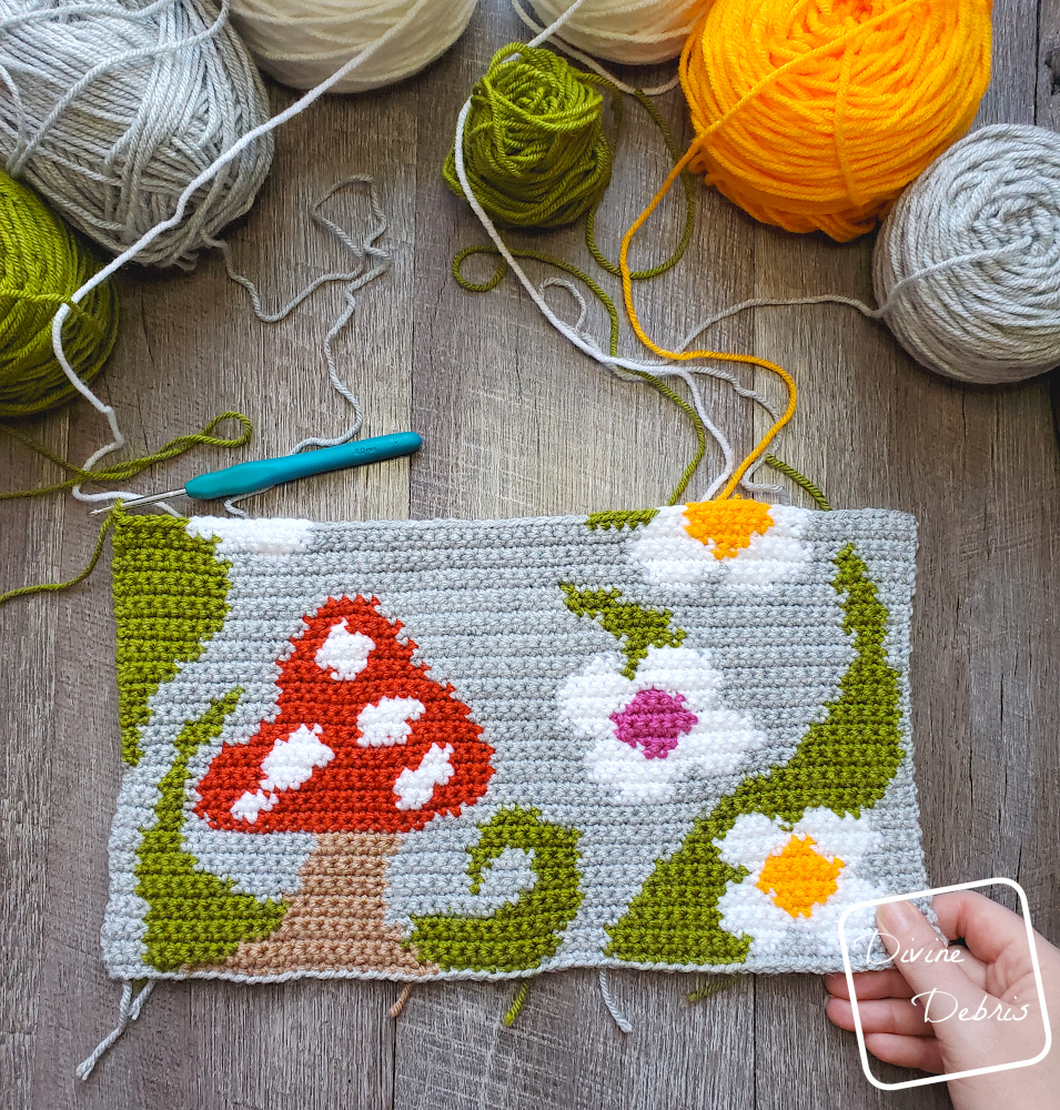 [Image description] A half finished 'Shrooms and Blooms Wall Hanging lays on a fake wood background with 7 cakes of yarn along the top of the photo and a white woman's hand holding the bottom right corner of the piece.