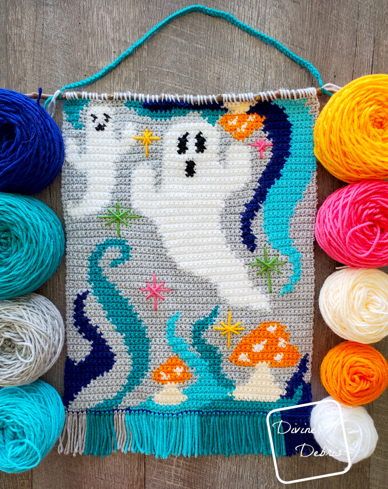 [Image description] the Ghosts of the Blue Lagoon Wall Hanging laying on a wood grain background surrounded by skeins of colorful yarn down both sides
