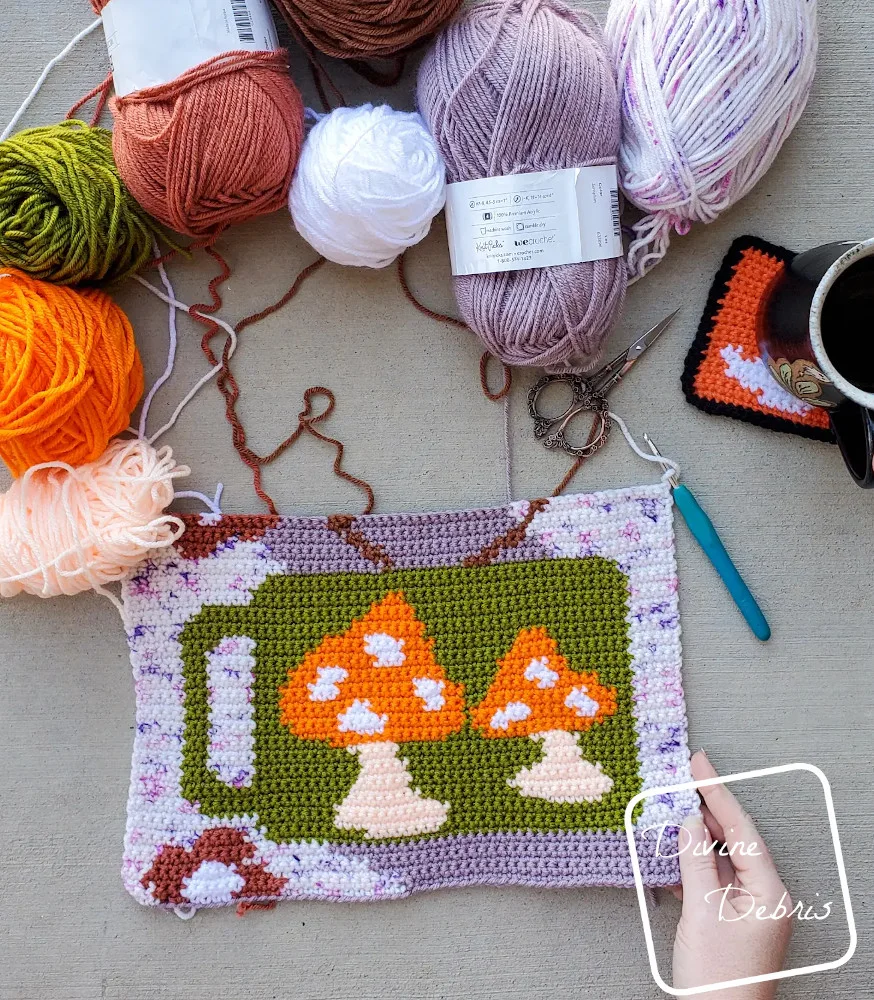 [Image description] A half finished Mug'Shroom Wall Hanging lays on cement while a white woman's hand holds the bottom right corner and the piece is surrounded by yarn skeins.