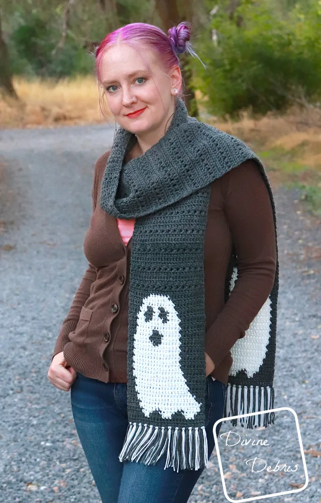 [Image description] A white woman with purple hair stands on a gravel trail in the woods, facing the camera wearing the Gloria Ghost Scarf from the crochet pattern, with only one end of the scarf showing.