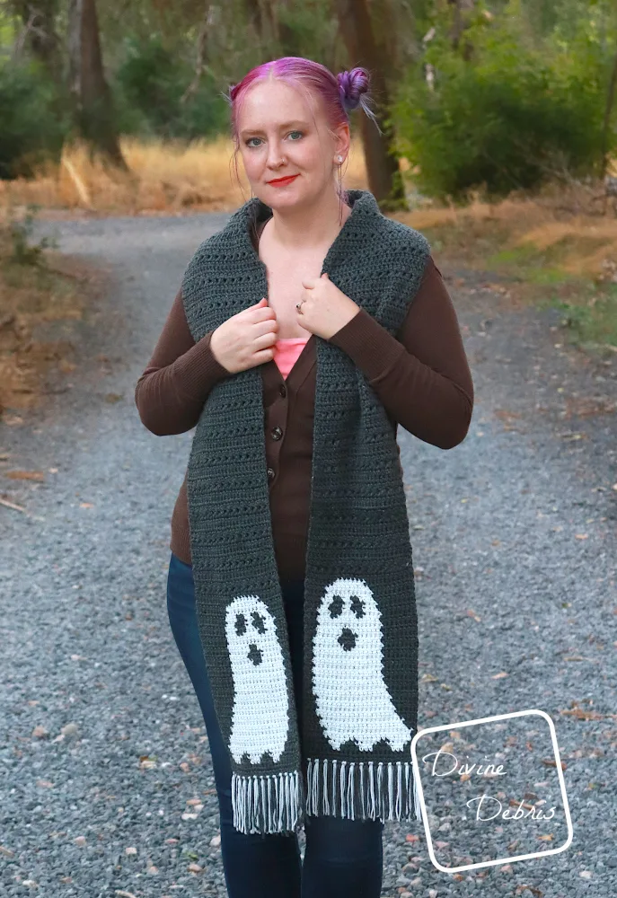 [Image description] A white woman with purple hair stands on a gravel trail in the woods, facing the camera wearing the Gloria Ghost Scarf crochet pattern, worn long.