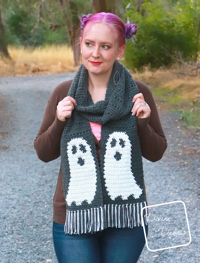 [Image description] A white woman with purple hair stands on a gravel trail in the woods, facing the camera looking to the left, wearing the Gloria Ghost Scarf from the crochet pattern