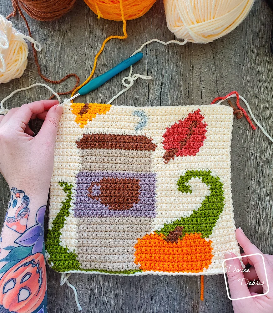 [Image description] A top down view of the partially finished Fall Coffee Wall Hanging on a wood grain background, with a white woman's hand holding the top corner, with skeins of yarn along the top of the piece