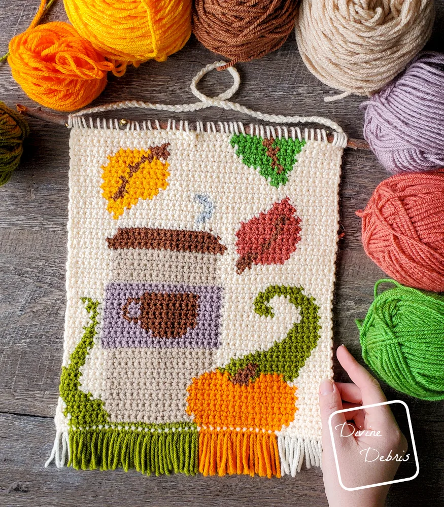 [Image description] A top down view of the finished Fall Coffee Wall Hanging on a wood grain background, with a white woman's hand holding the bottom right corner, with skeins of yarn along the top and side of the piece