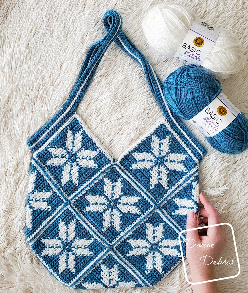 [image description] top down view of the Susie Snowflake Bag laying on a white faux fir background, with 2 skeins of yarn along the top of the photo and a white woman's hand holds the bottom right of the bag.