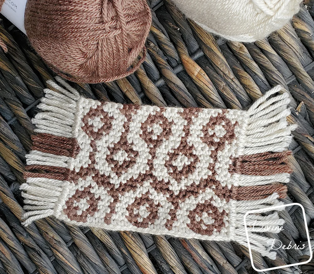 [Image description] Top down view of a single Torrance Mug Rug crochet coaster on a woven wood background and skeins of yarn along the top of the photo.