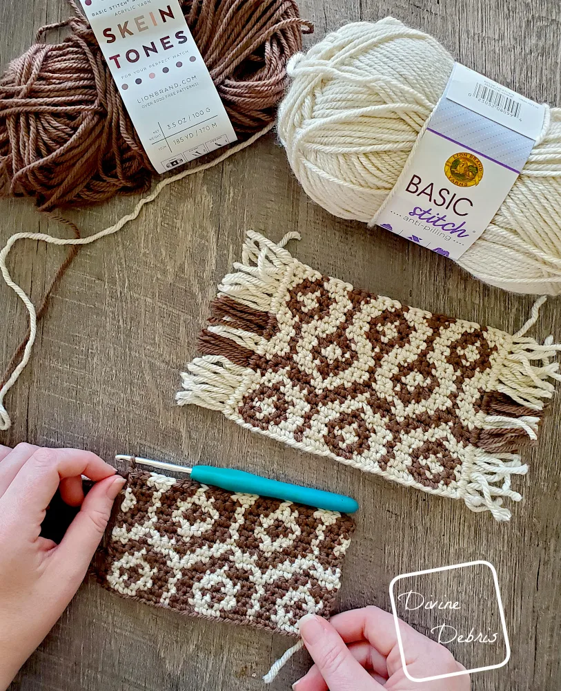 [Image description] Top down view of a Torrance Mug Rugs crochet coaster fake wood background, with unfinished mug rug in a white woman's hands, and skeins of yarn along the top of the photo.