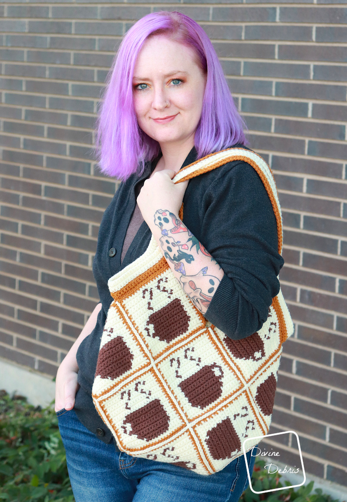 [image description] A white woman with purple hair stands against a brick background with the Calliope Coffee Bag hanging on her shoulder.