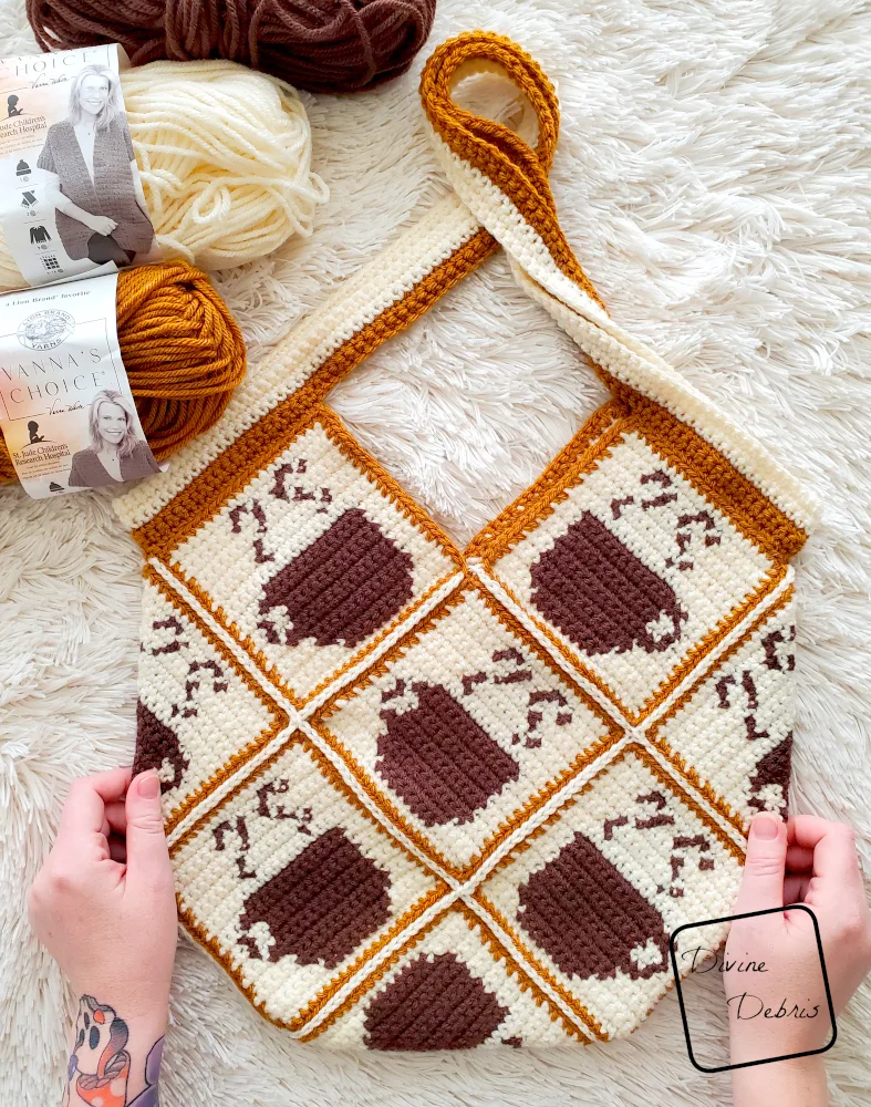 [Image description] Top down look of the Calliope Coffee Bag laying on a white faux fir blanket, with pair of white woman's hands holding the bottom of the bag and skeins of yarn along the top left corner.