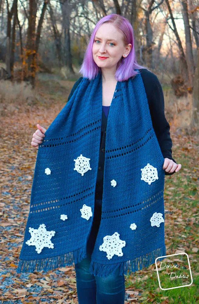 [Image description] A white woman with purple hair stands on a leaf covered trail in front of trees holding the Crystal Snowflakes Shawl on her shoulders and up to the viewer.