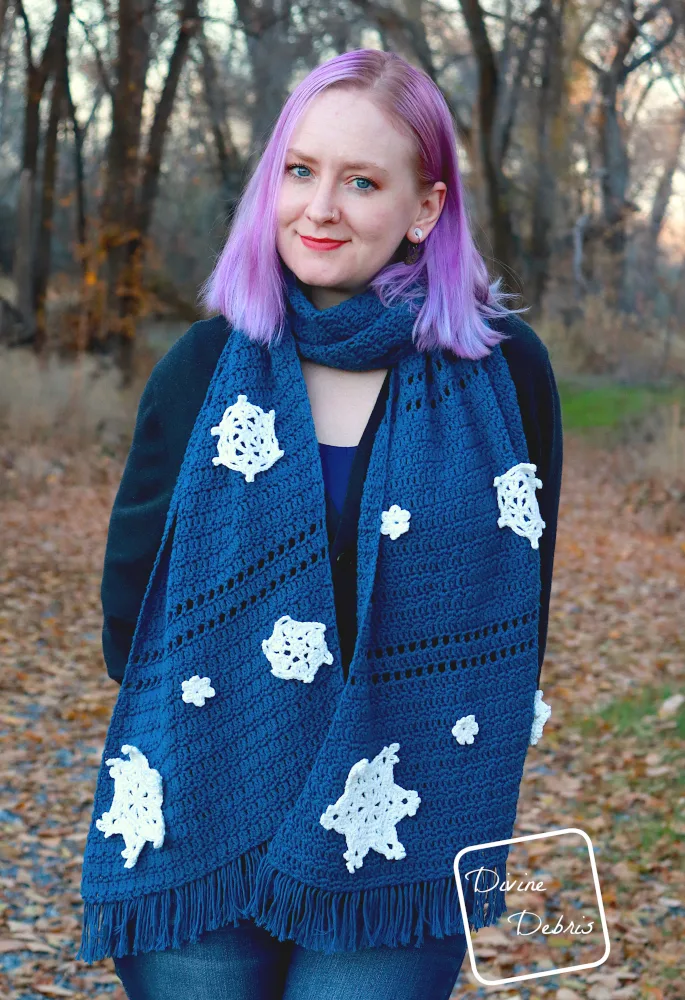 [Image description] A white woman with purple hair stands on a leaf covered trail in front of trees holding the Crystal Snowflakes Shawl around her neck and up to the viewer.