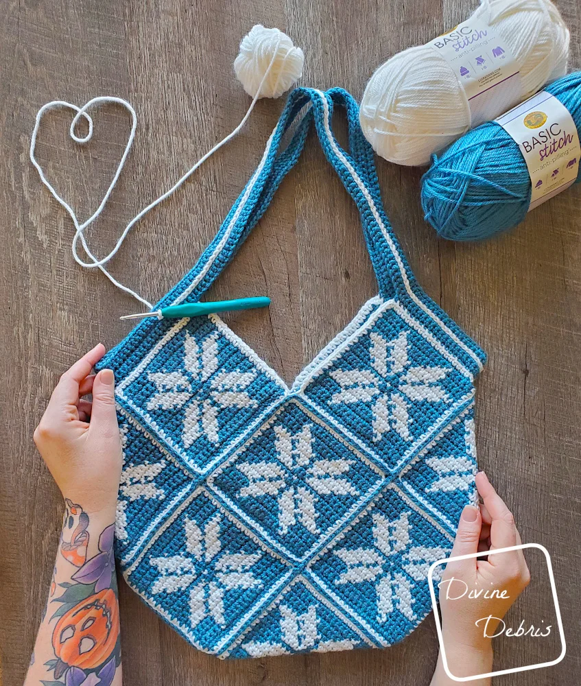 [image description] top down look of Susie Snowflake Bag with 2 skeins in the top right, a heart made from the yarn on the top left and a white woman's hands holding the bottom of the bag.