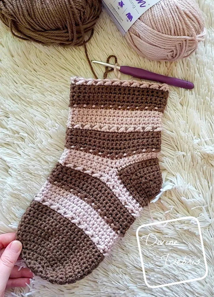[Image description] top down view of an unfinished Claire Stocking on a white fuzzy blanket, with 2 skeins of yarn along the top of the photo and a white woman's hand holding the toe.