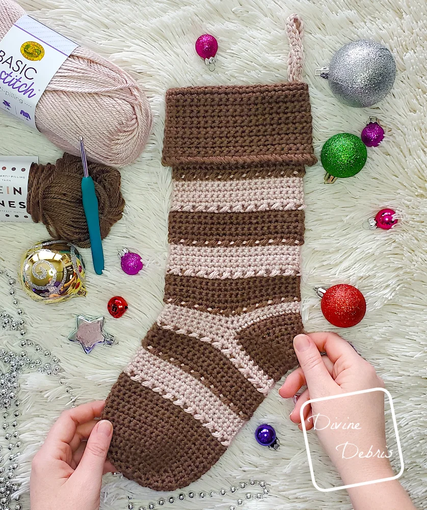 [image description] a top down view of the Claire Stocking laying on a white fuzzy blanket, surrounded by Christmas ornaments, with a white woman's hands holding the stocking by the heel and toe.