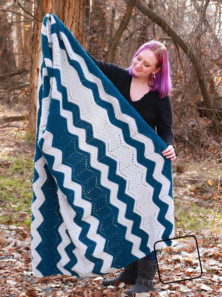 [image description] A white woman with purple hair stands in front of trees looking holding the Colorful Chevrons Blanket at an angle in front her.