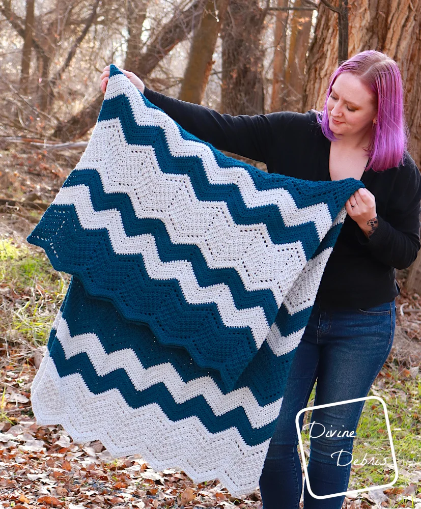 [image description] A white woman with purple hair stands in front of trees looking holding the Colorful Chevrons Blanket folded in half at an angle in front her.