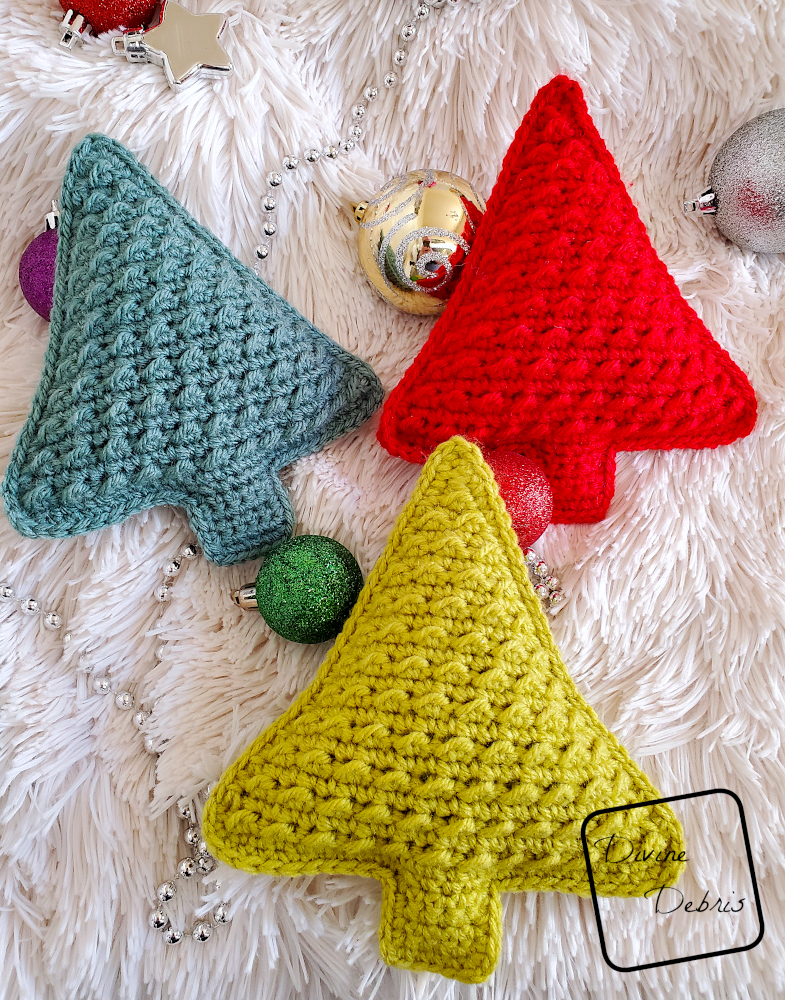 [Image description] 3 Cute Tree Amigurumis lay close together on a fuzzy white blanket with Christmas tree ornaments around them.