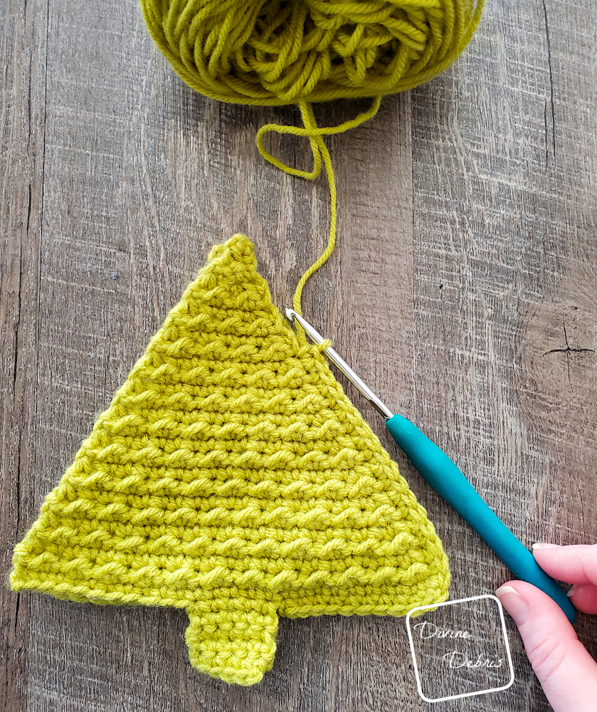 [Image description] Top down view of a Cute Tree Amigurumi on a wood grain background in lime green, in the process of seaming. A white woman's hand holds a crochet hook in the bottom right corner. 