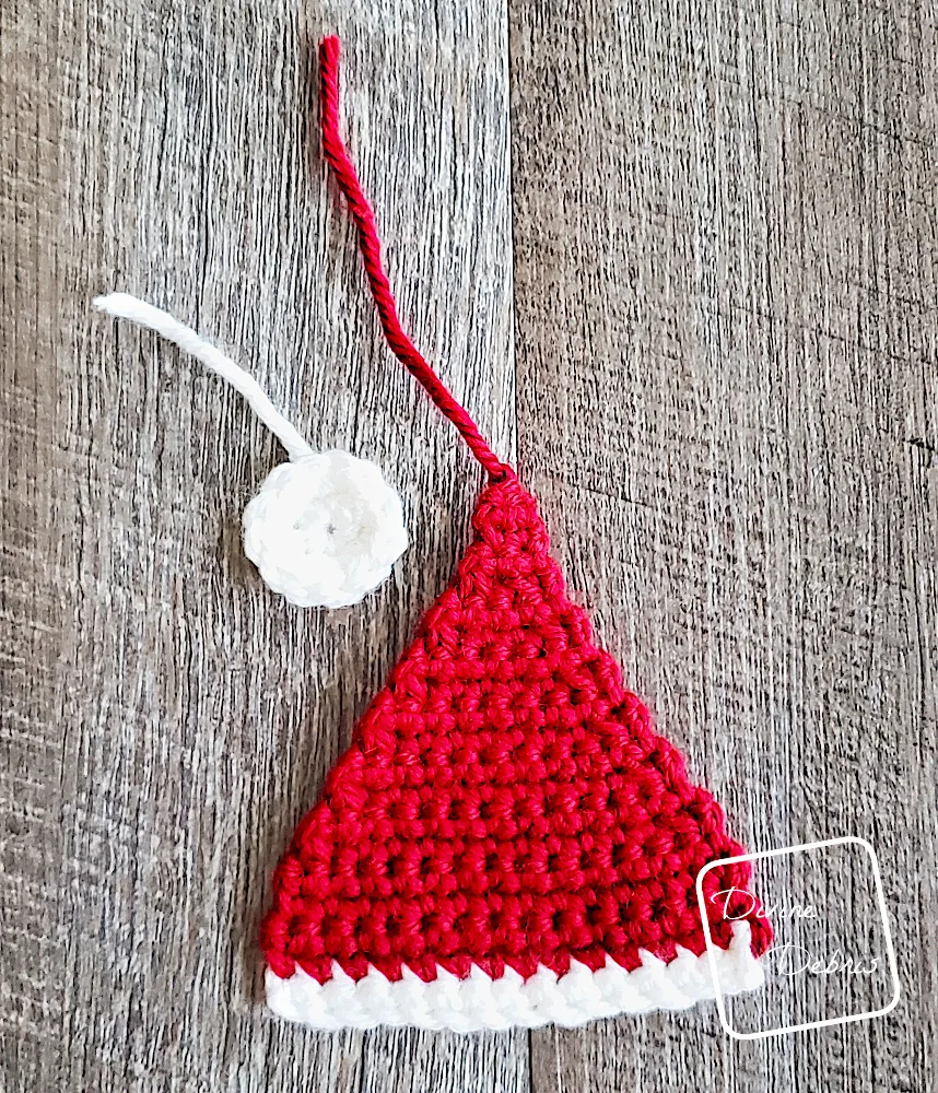 [Image description] Top down look at a unfinished Santa Hat Applique in red and a white pom pom laying on a fake wood background.