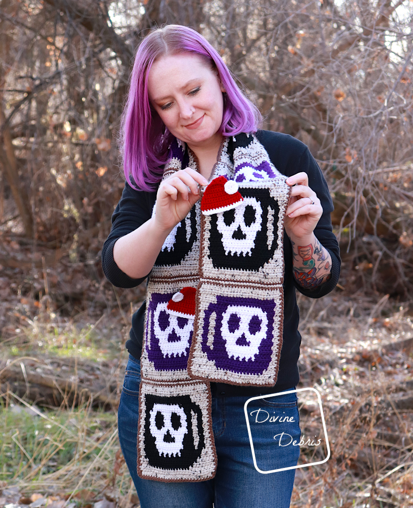 [Image description] A white woman with purple hair stands in front of trees holding a scarf up to the camera, with the Santa Hat Applique over the skull in the scarf design.