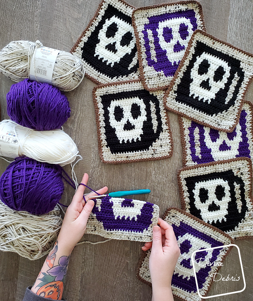[Image description] Top down look at a white woman's hands making a Wake the Dead square with skeins of yarn along the left side of the photo and finished Wake the Dead squares on the right.