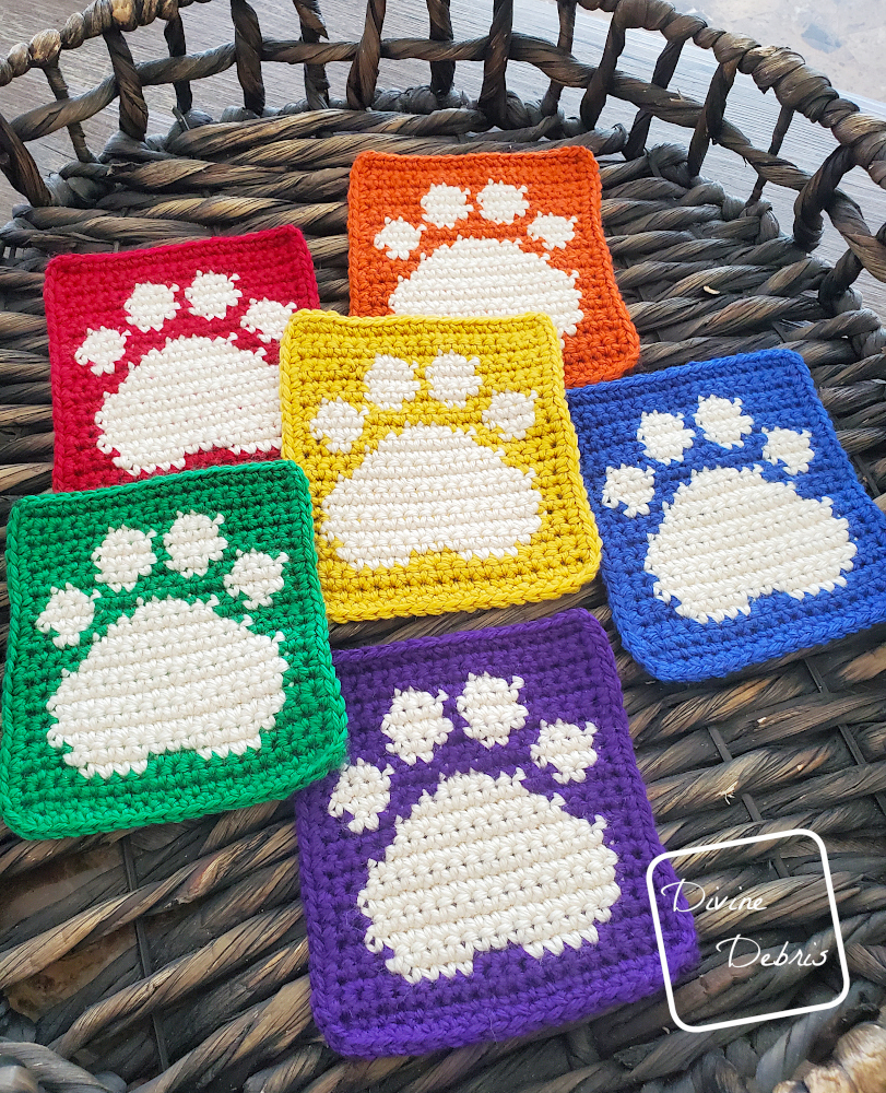 [Image description] A slightly angled view of 6 rainbow colored Cute Paw Print Squares in a wooden woven basket. 